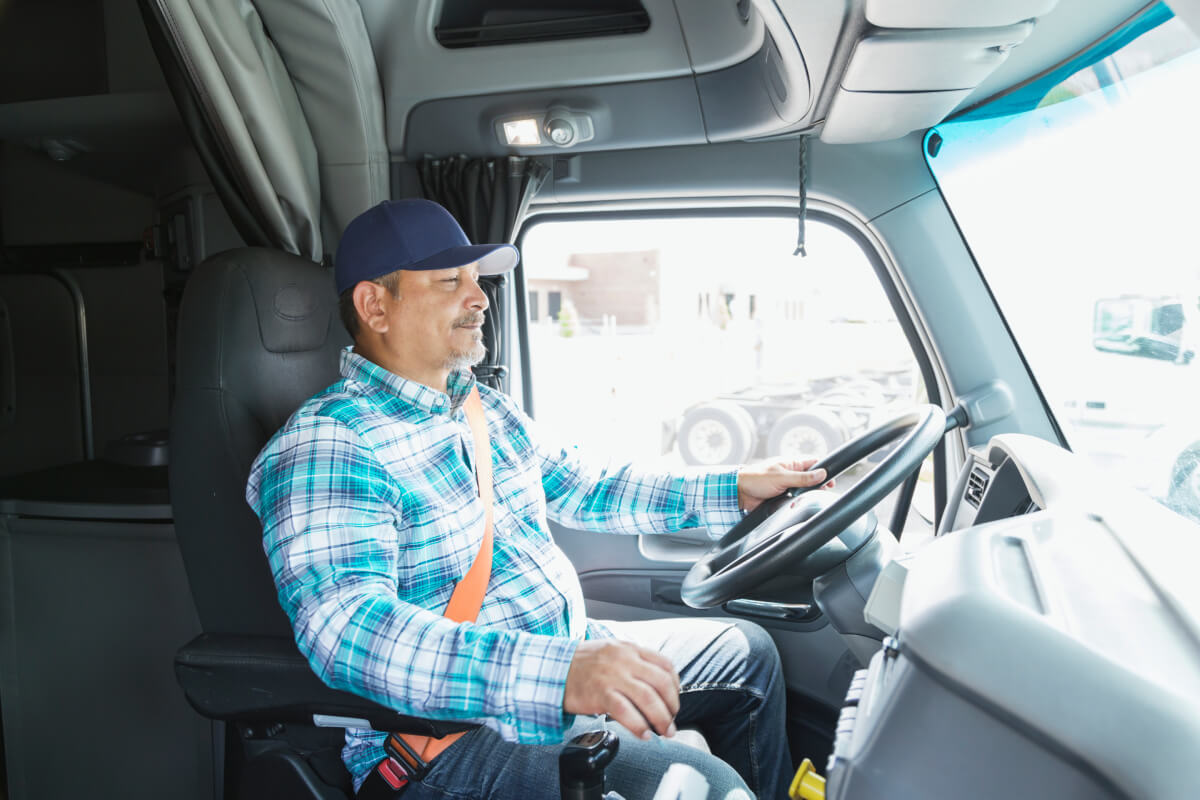 Mature Hispanic driver in a blue button-down shirt driving a semi-truck, to show the importance of owner operator insurance to protect against accident when operating your own trucking business.