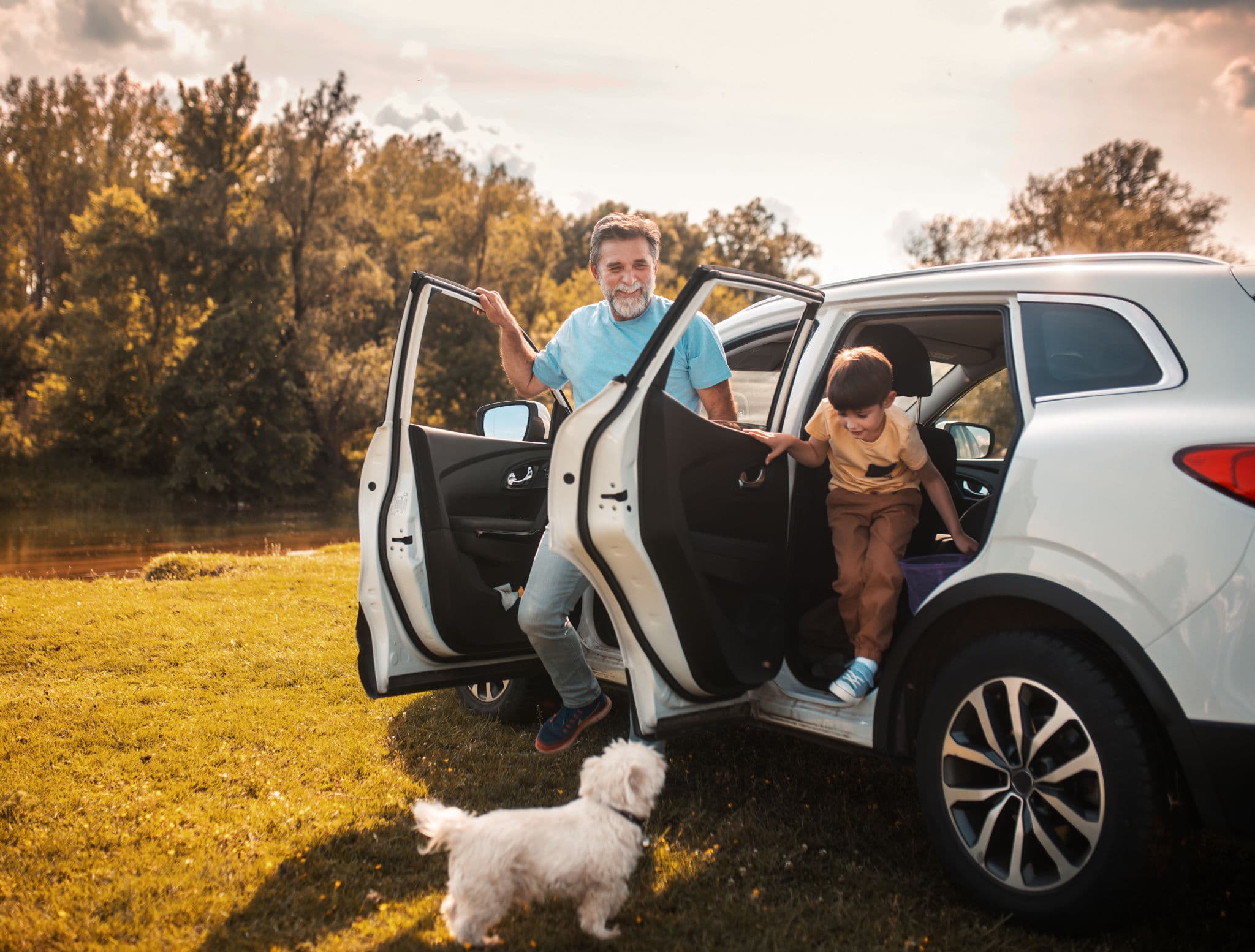 An older man and a young child step out of a white SUV on a sunny field, with a small white dog beside them