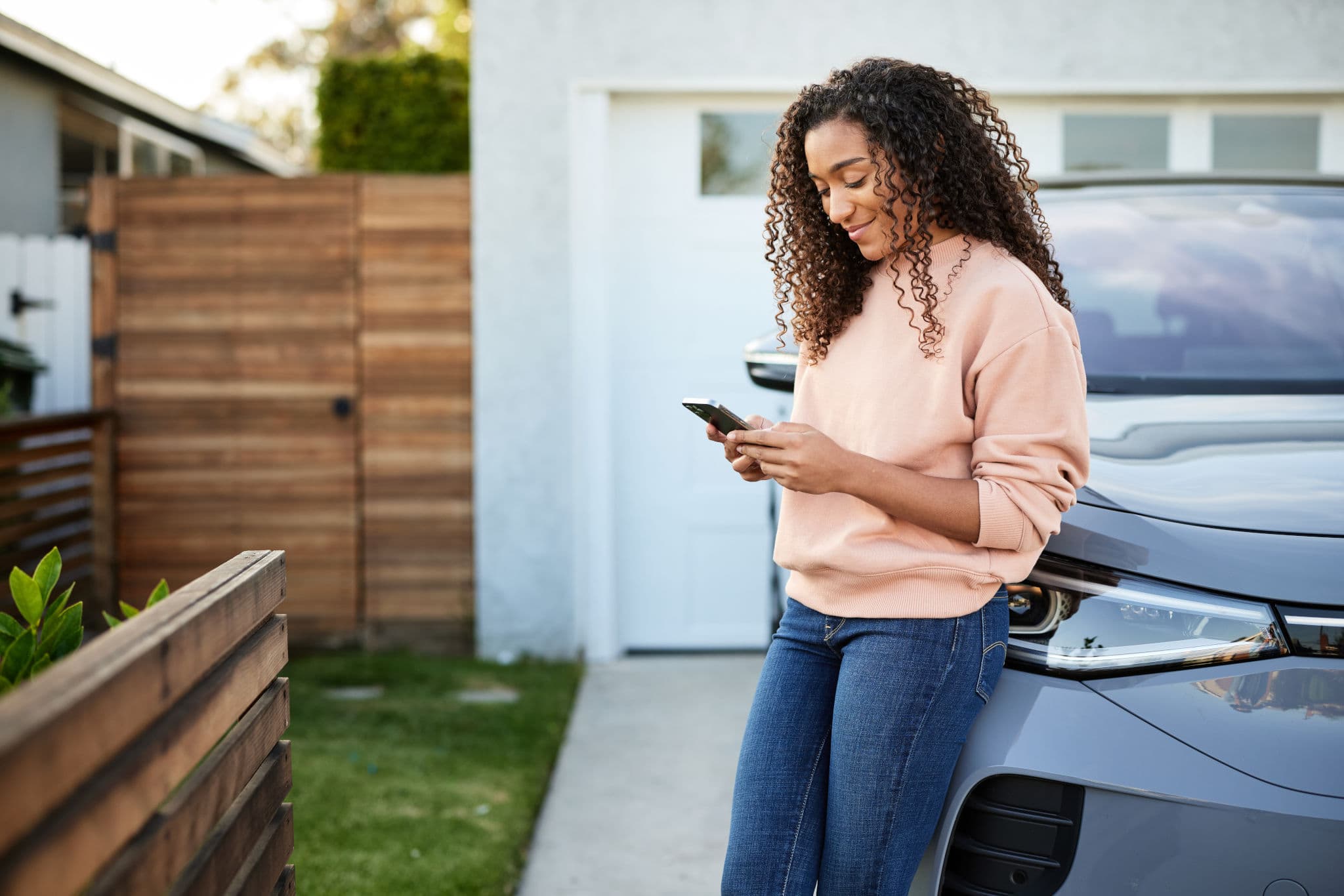 Woman leans on her car in the driveway and checks her phone, a convenient way to renew car insurance