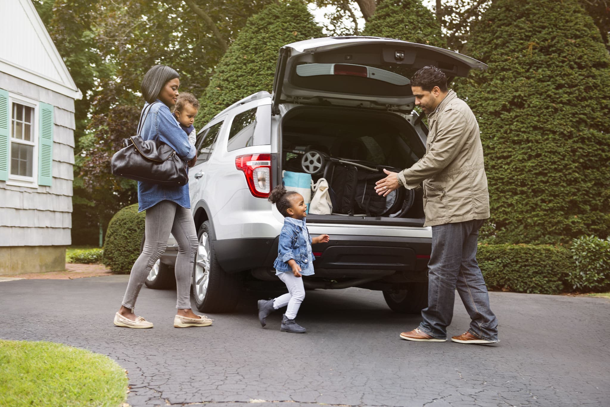 Parents and two small children load a white SUV in their driveway, a household that may need second car insurance