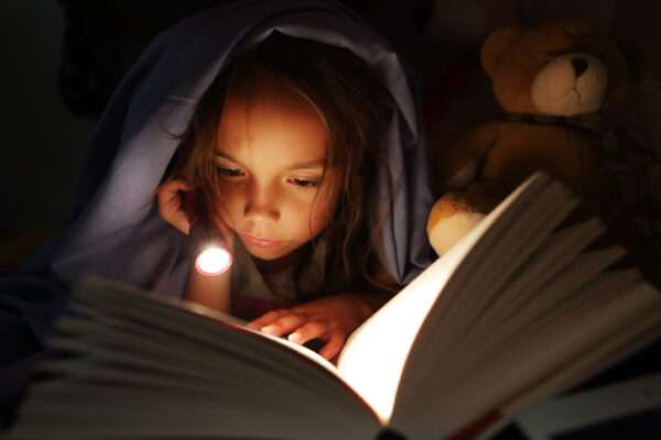 Girl under bed covers reading book by torchlight