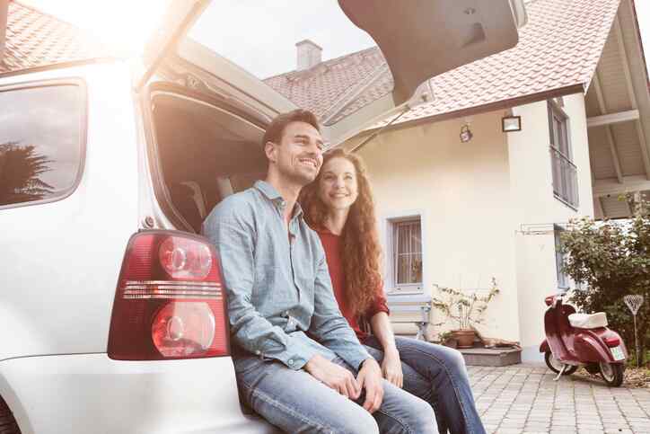 Smiling couple sitting in their car trunk on their driveway, with their house in the background and a red motorcycle nearby, showing how easy it is to bundle with GEICO Insurance Agency.