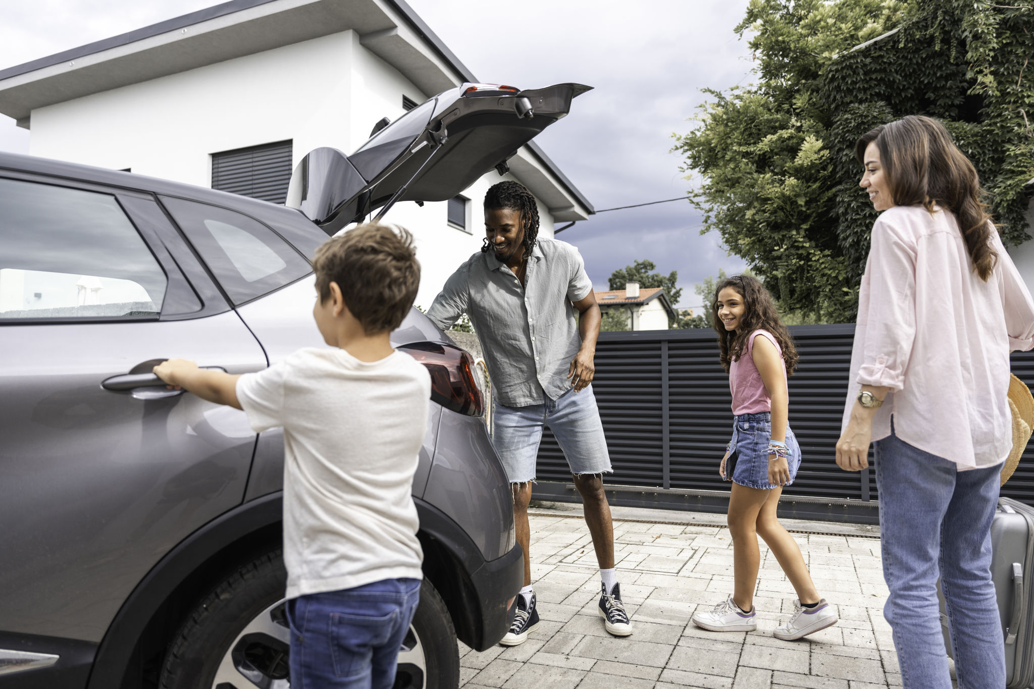 Two adults and two children gather around the open back of a gray SUV outside a home