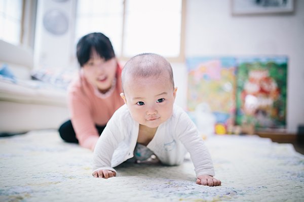 A baby crawling across the floor with their mom watching in the background.