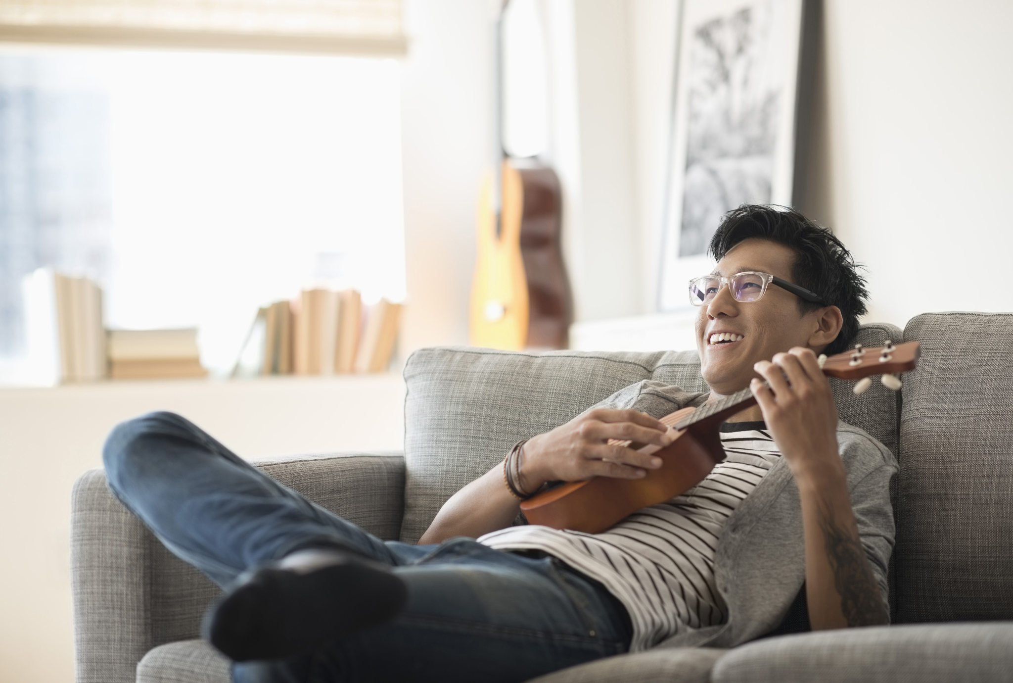 smiling renter relaxing on a gray couch playing a ukulele in his apartment, illustrating peace of mind basic renters insurance offers for your personal belongings