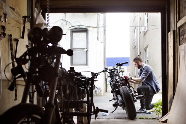 A man squatting next to his bike as he fixes it outside of his garage.