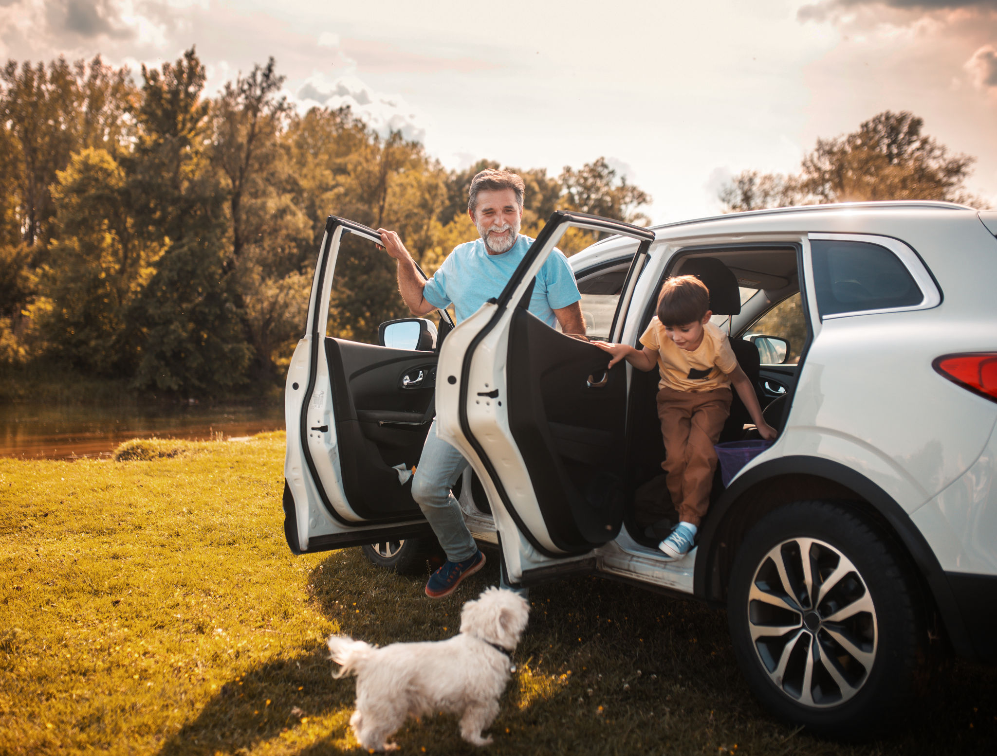 An older man and a young child step out of a white SUV on a sunny field, with a small white dog beside them