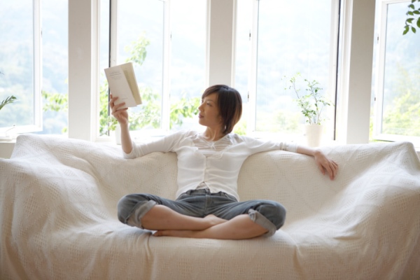 Woman sitting on a couch in front of a wall of windows and reading a book.