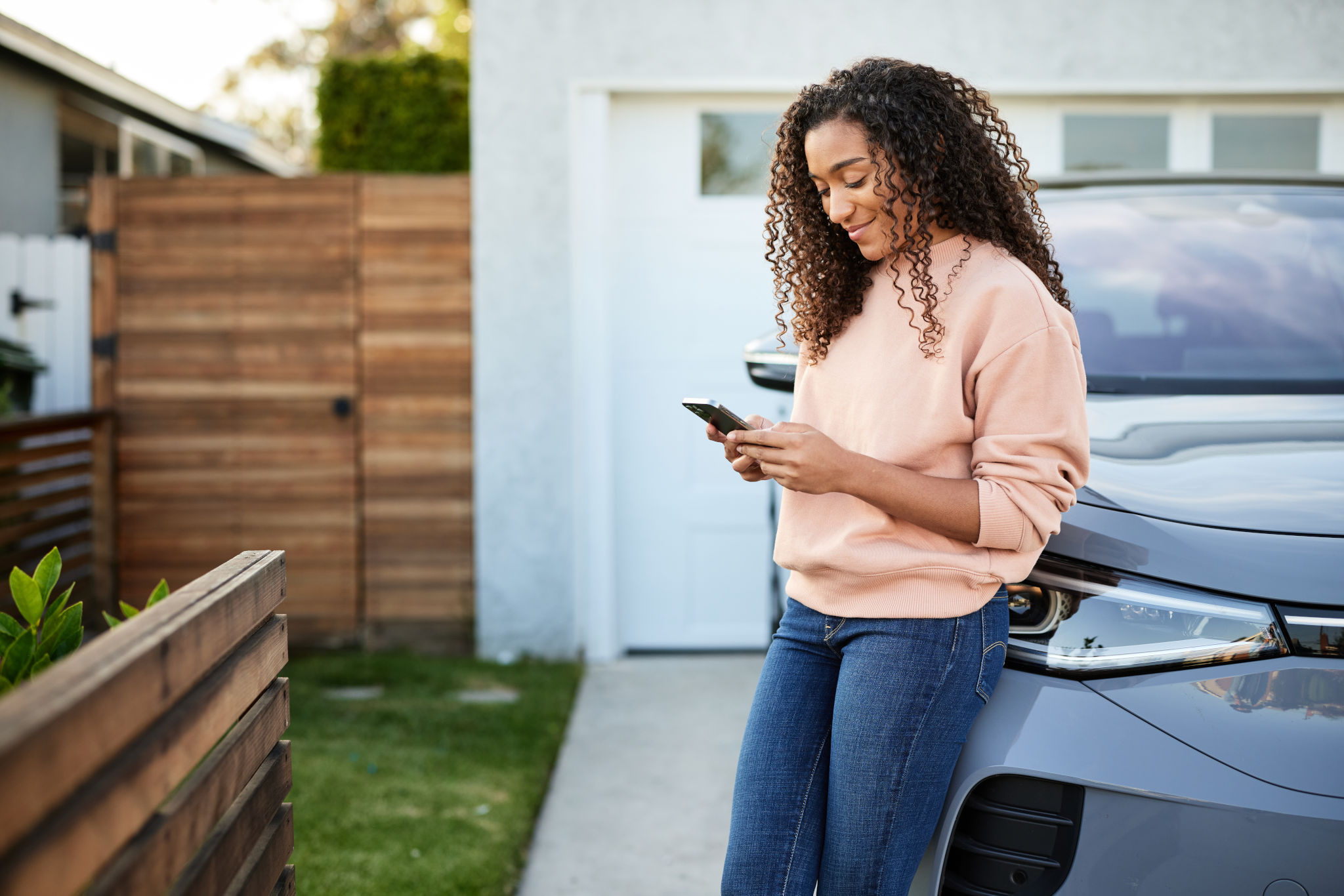 Woman leans on her car in the driveway and checks her phone, a convenient way to renew car insurance
