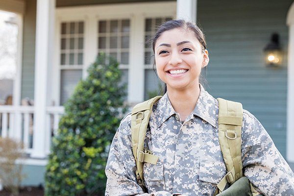 Woman dressed in military fatigues, smiling in front of a home.