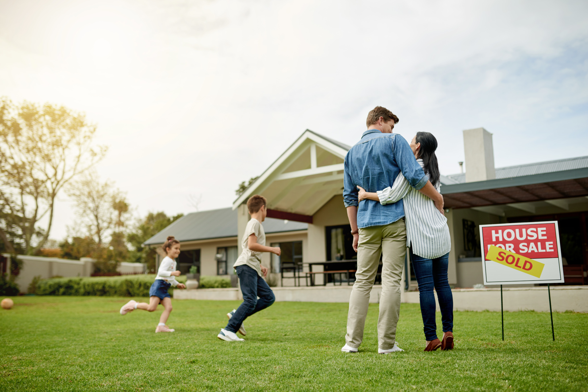 A couple embrace on the lawn as two children run toward a house with a sold sign
