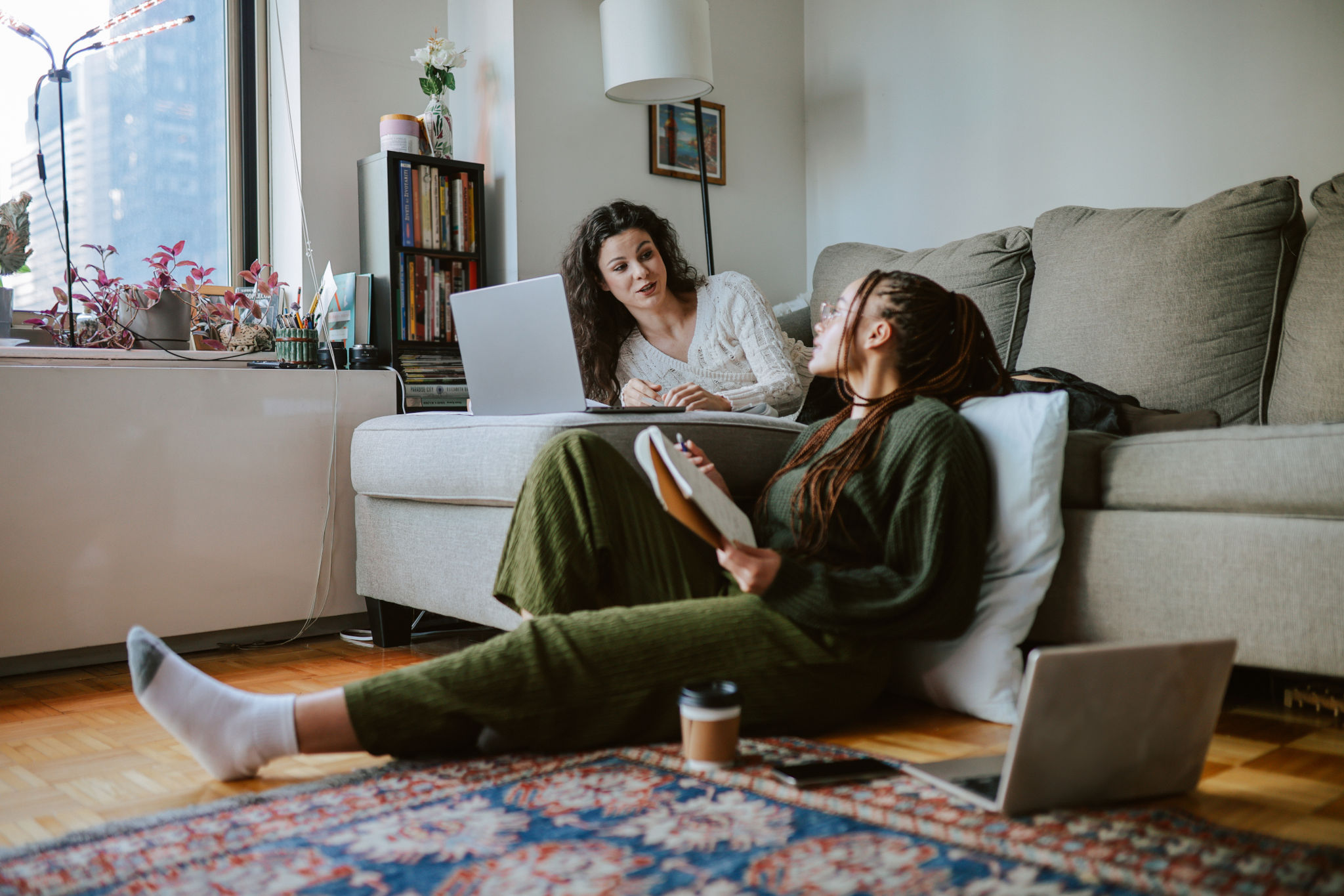 Two roommates talk in their living room, one sitting on the floor with a notebook