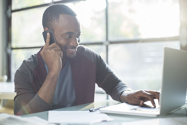 Man talking on the phone and smiling while typing on his laptop.