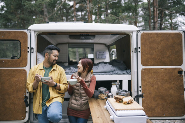 A couple enjoying a meal behind their camping van that is set up with all the doors open.